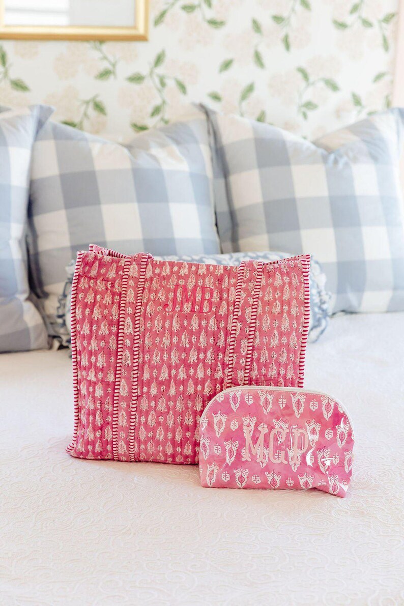 a pink and white patterned bag and a matching pink makeup bag placed on a bed with a floral wallpaper.