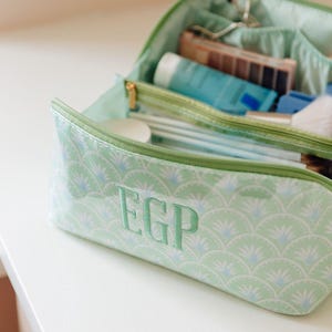 May include: A light green cosmetic bag with a repeating white and blue fan-shaped pattern and the embroidered letters EGP on the front. The bag features a zippered closure and an open top compartment containing various beauty and grooming items.