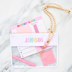 Peut inclure: Une pochette en vinyle transparent avec une bordure blanche et le nom JOHNSON brodé en fil arc-en-ciel. À l'intérieur se trouvent un bloc-notes hebdomadaire, des notes adhésives roses et un stylo rose. Un cordon perlé avec une breloque pêche.
