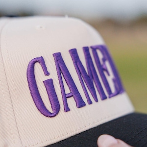May include: A beige baseball cap with the word "GAME" embroidered in purple. The cap has a black brim and a classic, structured design. The text is in a bold, stylized font.