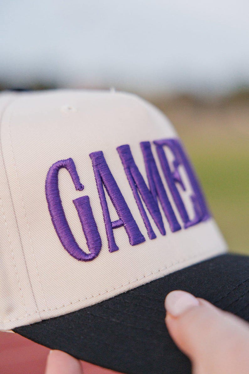 May include: A beige baseball cap with the word "GAME" embroidered in purple. The cap has a black brim and a classic, structured design. The text is in a bold, stylized font.