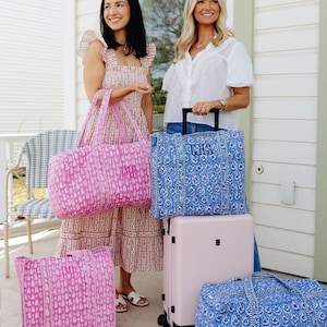 Two women standing on a porch, each holding a large bag, with a pink and blue patterned bag between them.