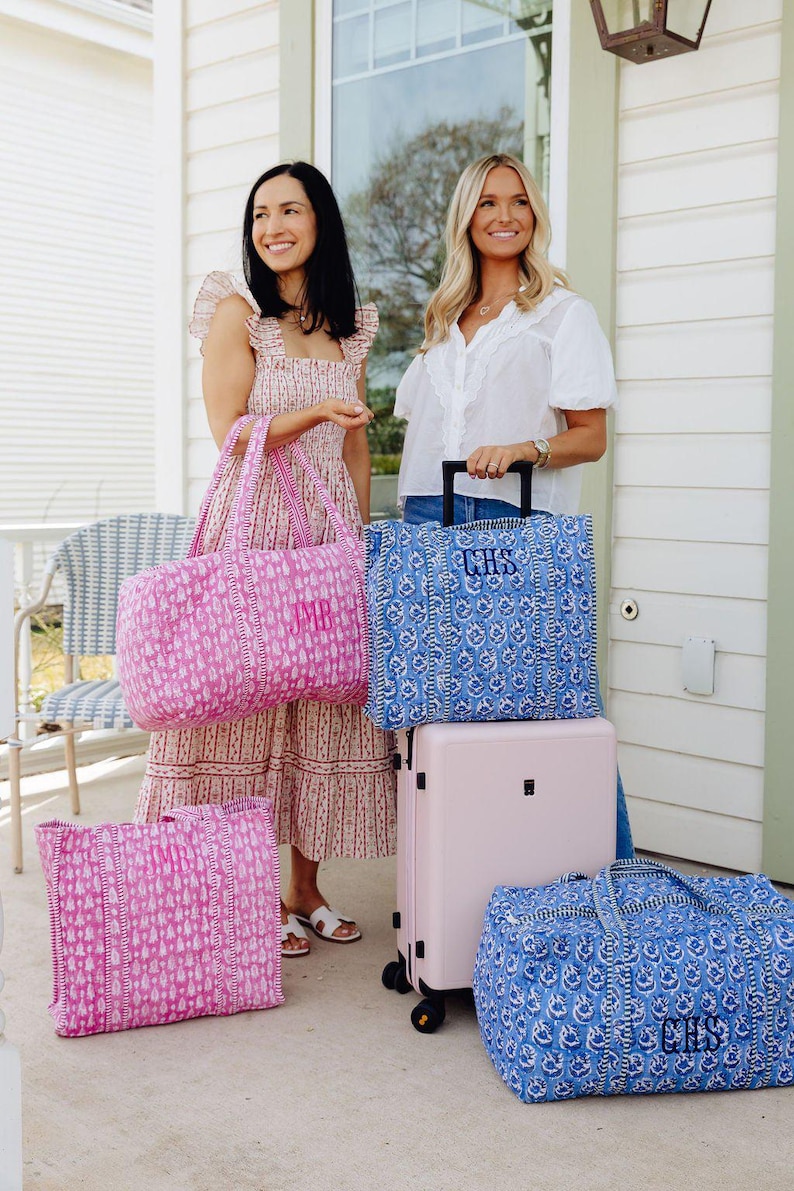 Two women standing on a porch, each holding a large bag, with a pink and blue patterned bag between them.
