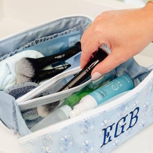 May include: A light blue rectangular cosmetic bag with a white floral pattern and the embroidered monogram EGB in dark blue. The open bag contains various makeup brushes, a hair brush, and a bottle of e.l.f. Holy Hydration serum inside.