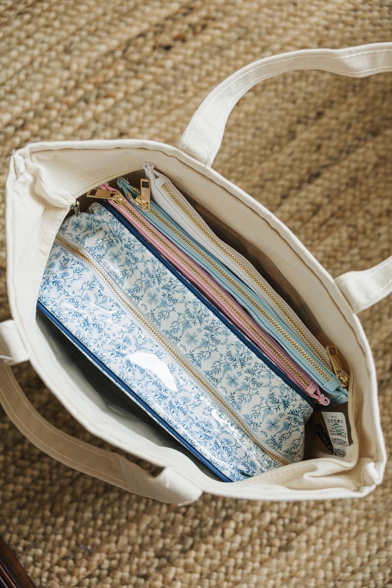 May include: A cream-colored tote bag holds several zippered pouches. The pouches feature floral patterns in blue and white, with gold zippers. Additional pouches are solid colors: pink, light blue, and white.