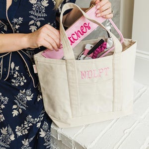 May include: A cream-colored tote bag with the embroidered letters "NOLPT" on the front pocket. Inside, a pink zippered pouch with "stitches" in pink script is visible, along with other sewing supplies. The bag has sturdy handles and a rectangular shape.