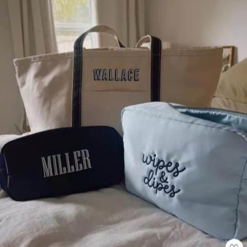 May include: A large beige canvas tote bag with dark blue handles and the name WALLACE embroidered in blue. In front are two smaller bags: a black pouch with the name MILLER embroidered in white and a light blue pouch with wipes & dipes embroidered.