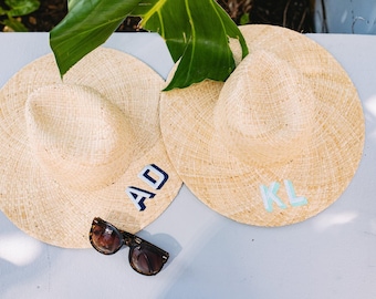 May include: Two straw sun hats with initials "AD" and "KL" embroidered on them. The hats are on a white surface with a green leaf in the background.