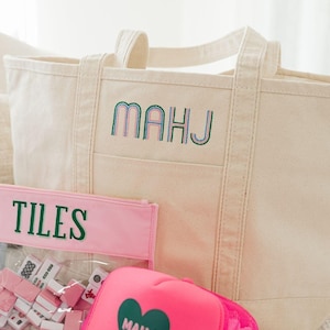 May include: A cream-colored canvas tote bag with the embroidered text MAHJ, a clear pink zippered pouch containing white and pink game tiles labeled TILES, and a bright pink trucker hat featuring a green heart with the text MAHJ.