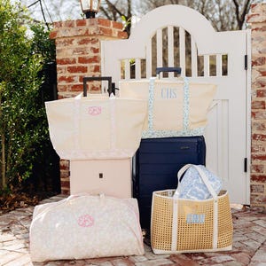 May include: A collection of travel bags and luggage in various sizes and styles. The bags are cream-colored with floral patterns and monogrammed initials. A navy blue suitcase and a woven tote bag are also included.