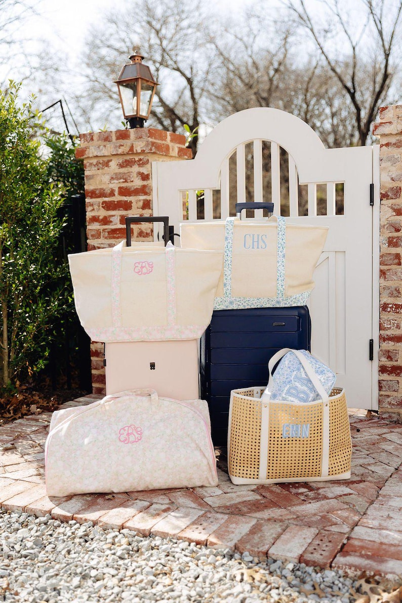 May include: A collection of travel bags and luggage in various sizes and styles. The bags are cream-colored with floral patterns and monogrammed initials. A navy blue suitcase and a woven tote bag are also included.