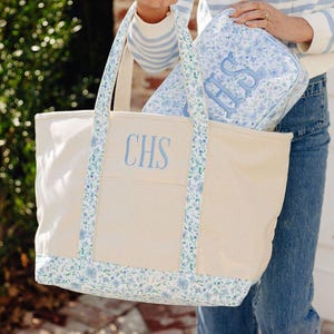 May include: A canvas tote bag with blue floral trim and the embroidered initials "CHS" in light blue. A matching floral print cosmetic bag with the initials "HS" is inside the tote. The bag has a cream-colored body and a rectangular shape.