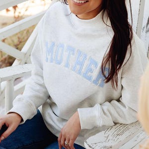 May include: A light gray heathered sweatshirt featuring a mock neck collar and long sleeves. The word MOTHER is appliqued across the chest in light blue block letters with a white outline. The garment is paired with dark blue denim jeans.