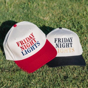 Könnte beinhalten: Zwei Baseballkappen mit dem Text "FRIDAY NIGHT LIGHTS" auf der Vorderseite gestickt. Eine Kappe hat eine weiße Krone und einen roten Schirm, die andere hat eine weiße Krone und einen schwarzen Schirm. Beide liegen auf grünem Gras.