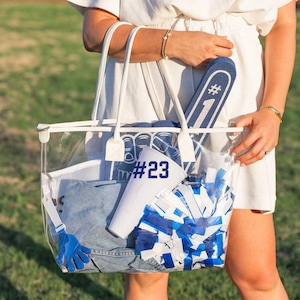 May include: A clear tote bag filled with blue and white pom-poms, a denim garment, a white megaphone, and a blue foam finger. The bag also contains a white towel and a blue glove. The text "#23" is visible on a white item.