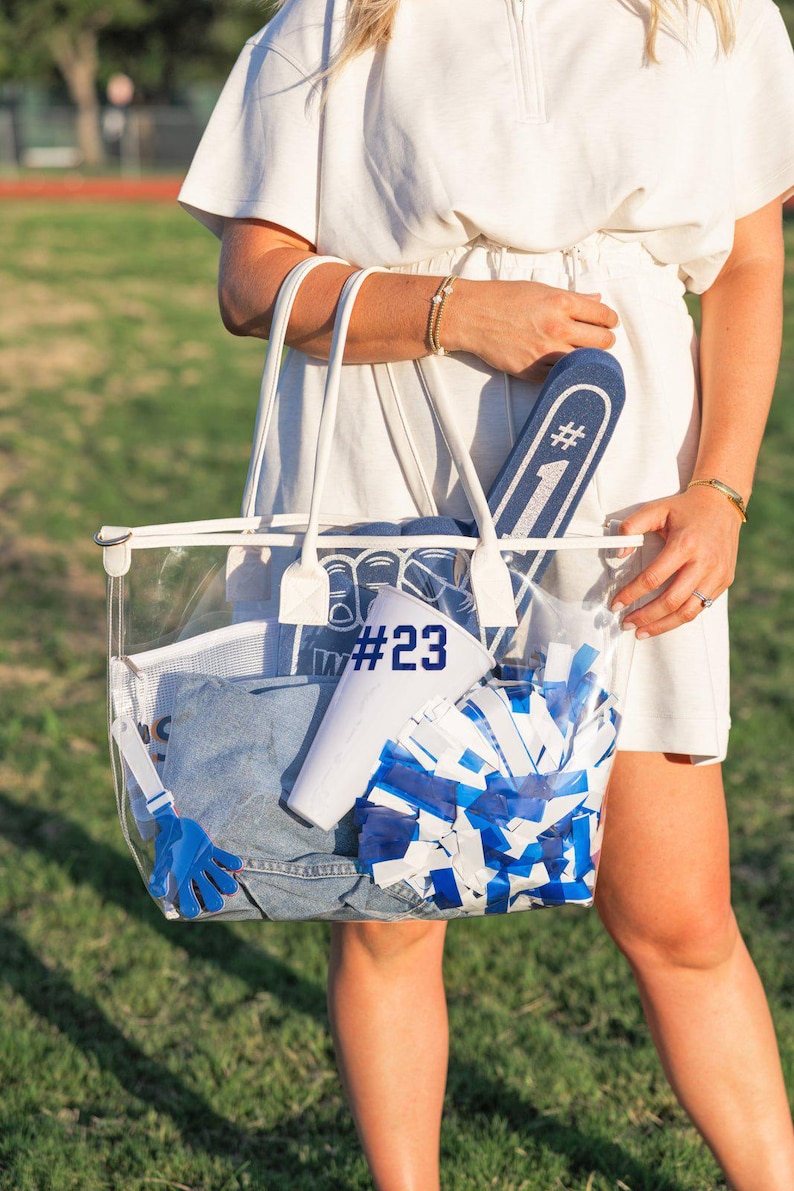 May include: A clear tote bag filled with blue and white pom-poms, a denim garment, a white megaphone, and a blue foam finger. The bag also contains a white towel and a blue glove. The text "#23" is visible on a white item.