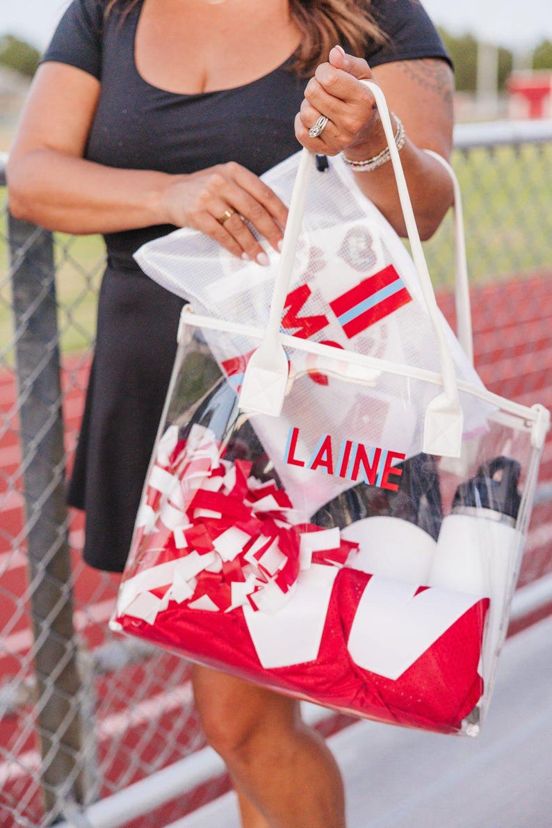 May include: Clear tote bag filled with red and white items, including a pom-pom, jersey, and water bottle. The name "LAINE" is printed on the front in red and blue letters. The bag has white handles and is held by a person.
