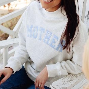 May include: A light gray heathered sweatshirt featuring a mock neck collar and long sleeves. The word MOTHER is embroidered across the chest in light blue block letters with a white outline, creating a bold and clear graphic design.