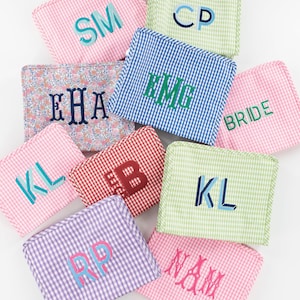May include: A collection of rectangular zippered pouches in various gingham and floral patterns, each featuring personalized embroidered monograms or text including SM, CP, EHA, HMG, BRIDE, KL, B, KL, RP, and NAM in diverse thread colors.