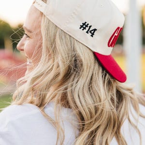 May include: Cream-colored baseball cap with a red brim. The cap features the number "#14" in black on the side and a red "C" logo. The cap is worn by a person with blonde hair.
