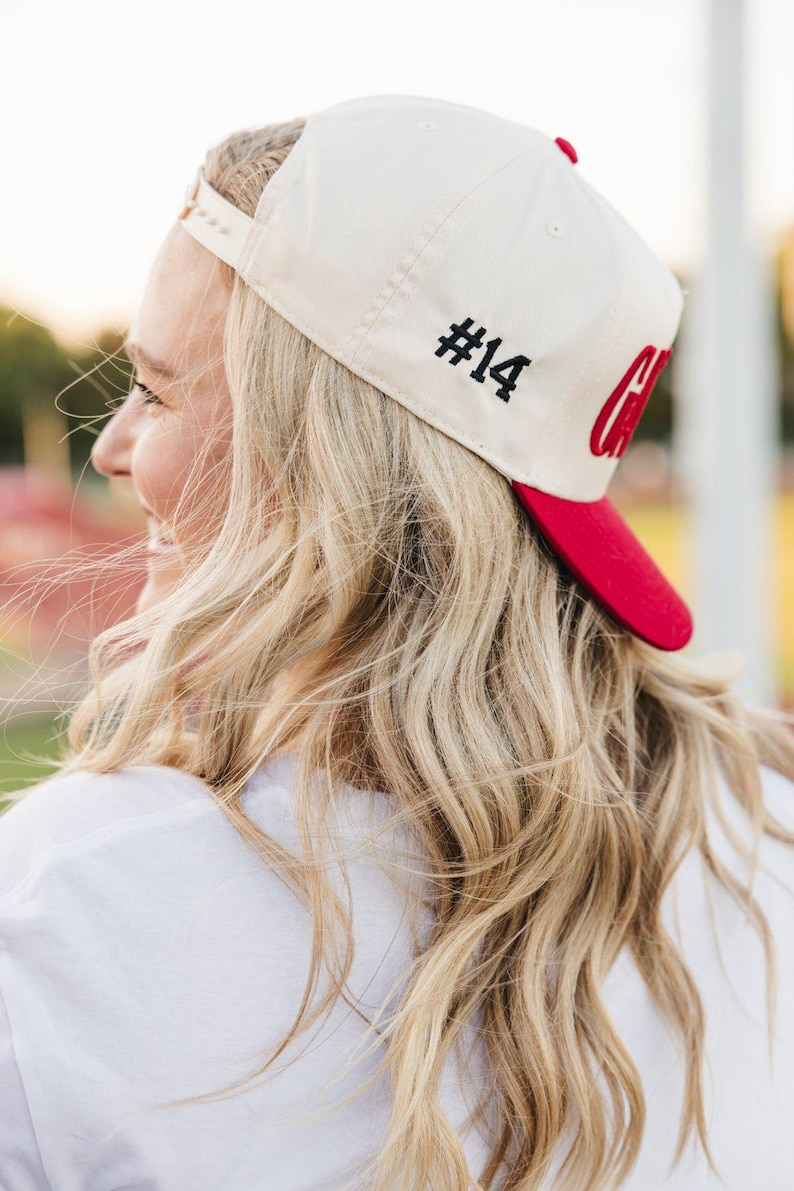 May include: Cream-colored baseball cap with a red brim. The cap features the number "#14" in black on the side and a red "C" logo. The cap is worn by a person with blonde hair.