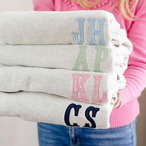 May include: A stack of four light grey heather sweatshirts, each featuring two-letter embroidered monograms in different colours: light blue JH, light green AP, pink KL, and navy blue CS. The embroidery uses a classic serif font style.
