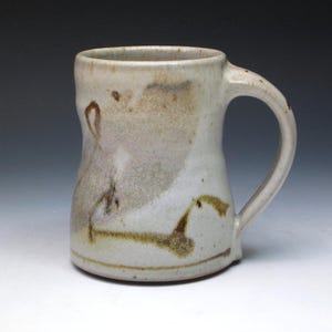May include: Handmade ceramic mug with a cream-colored base and a curved handle. The mug features brown and tan abstract designs and a slightly irregular shape, giving it a rustic appearance. The mug is suitable for hot or cold beverages.