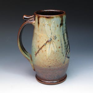 May include: A handmade ceramic mug with a dragonfly design. The mug features a tall, cylindrical shape with a large handle and a small thumb rest. The exterior has a mottled glaze in shades of brown, green, and tan. The dragonfly is etched into the surface.