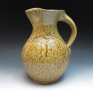 May include: A ceramic pitcher with a brown glaze and a design of a lizard. The pitcher has a wide mouth and a handle.