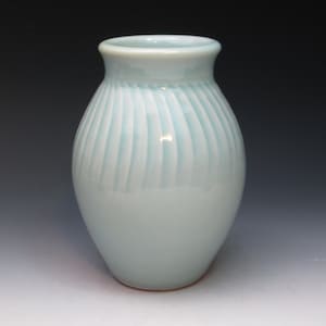 May include: A light blue ceramic vase with a textured, vertical striped pattern. The vase has a rounded body and a slightly flared rim. The vase is approximately 7 inches tall.