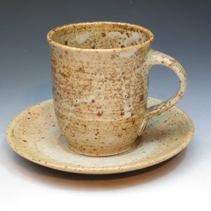 May include: A speckled beige ceramic coffee mug with a handle, sitting on a matching saucer.