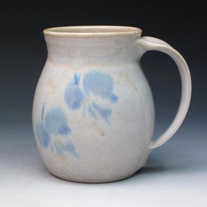 May include: A ceramic pitcher with a rounded body and a curved handle. The pitcher is white with light blue floral designs and a beige rim. The design is simple and elegant, suitable for serving beverages. The pitcher is approximately 15 cm tall.
