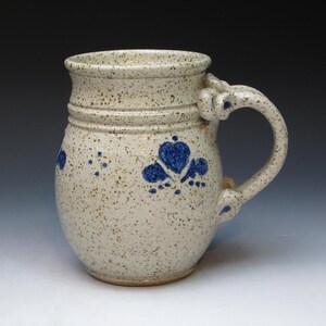 May include: A white ceramic mug with a speckled glaze and blue floral designs. The mug has a rounded body and a handle with a unique shape.