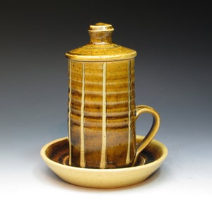 May include: A brown and yellow ceramic mug with a lid and a matching saucer. The mug has a striped pattern and a handle.
