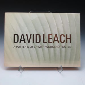 May include: A book titled "DAVID LEACH" with the subtitle "A POTTER'S LIFE WITH WORKSHOP NOTES." The book has a light beige cover with a subtle, textured pattern. The title is in a dark, reddish-brown font. A black handle is visible at the top.