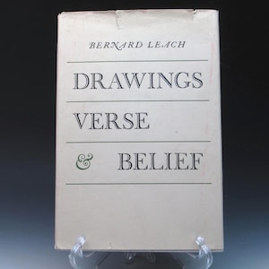 May include: A vintage book titled "DRAWINGS VERSE & BELIEF" by Bernard Leach. The book cover is a light beige color with black text and green accents. The book is displayed on a clear plastic stand.