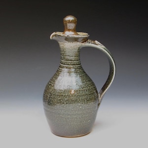 May include: A green ceramic pitcher with a lid and a handle. The pitcher has a speckled glaze and a brown rim.