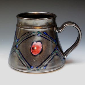 May include: A handmade ceramic mug with a dark metallic glaze. The mug features a red and pink central design, framed by blue and green accents. The handle is curved, and the mug has a wide base.