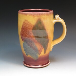 May include: A yellow ceramic mug with a brown, green, and blue glaze pattern. The mug has a handle with a small knob on the top.