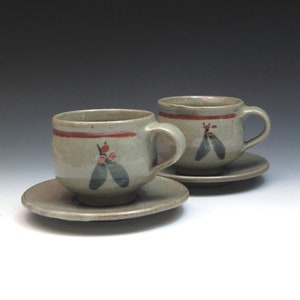 May include: Two ceramic teacups and saucers with a light green glaze and red and black painted designs. The cups have a rounded shape and a handle on the side. The saucers are round and have a slightly raised edge.