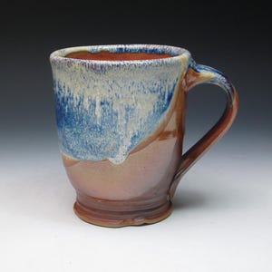 May include: A handcrafted ceramic mug with a distinctive glaze. The mug has a blue and white top that fades into a tan and brown base. The interior is a warm brown. The handle is a matching brown colour.