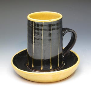 May include: A black ceramic mug with a yellow rim and vertical yellow stripes. The mug has a handle and sits on a matching saucer.