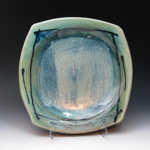 May include: A square ceramic serving dish with rounded corners. The dish has a light teal rim and a blue interior with a textured, speckled appearance. Black lines and drips decorate the rim. The dish is displayed on a clear stand.