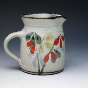 May include: A white ceramic pitcher with a brown rim and a floral design in red, yellow, and green. The pitcher has a handle and a rounded body.