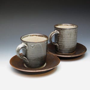 May include: Two ceramic coffee cups with saucers. The cups are a dark gray with a speckled glaze. The saucers are a dark brown.