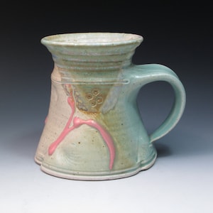 May include: A ceramic mug with a light green glaze and pink accents. The mug has a wide, flared rim and a handle. The glaze has a speckled appearance and there are small, raised dots on the side of the mug.