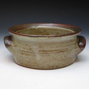 May include: A handmade ceramic bowl with two handles. The bowl has a light beige body with brown speckles and a dark brown rim. The handles are also dark brown. The bowl is suitable for serving soups, stews, or other dishes.
