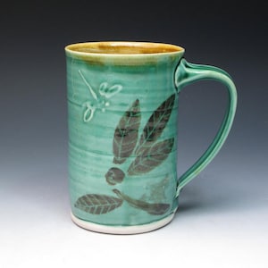 May include: A green ceramic mug with a brown leafy design and a dragonfly. The mug has a white rim and a handle.