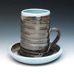 May include: A handmade ceramic mug with a matching saucer. The mug is cylindrical with a dark gray and brown striped pattern, a curved handle, and a light blue rim. The saucer is light blue.