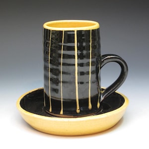 May include: A black ceramic mug with yellow stripes and a matching saucer. The mug has a handle and is sitting on the saucer.